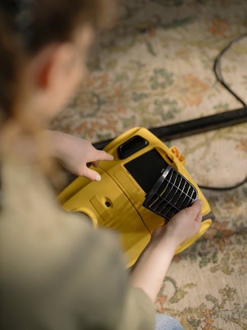 A person operating a yellow portable carpet cleaning machine, with a black vent grille, on an ornate patterned area rug in a well-lit room. The person's hands are visible, holding the machine's handle