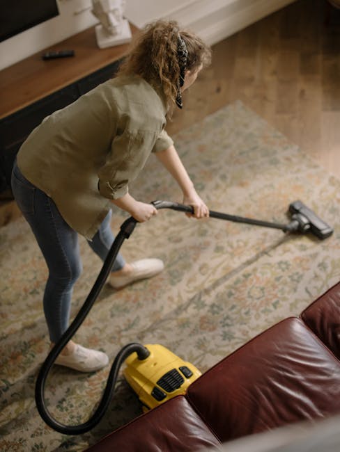 A professional cleaner dressed in a white protective suit, face mask, and gloves is performing deep cleaning on a beige carpet in a well-lit living room. The cleaner is using a cleaning machine to ext