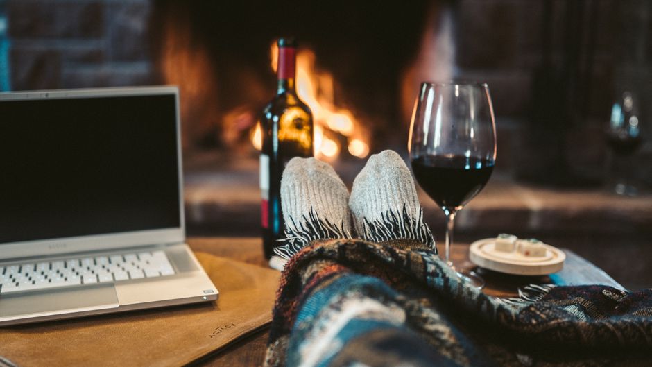 A person relaxing by a fireplace in a living room, with their feet up on a wooden coffee table covered by a colorful blanket. On the table, there is a closed silver laptop, a glass of red wine, a bott